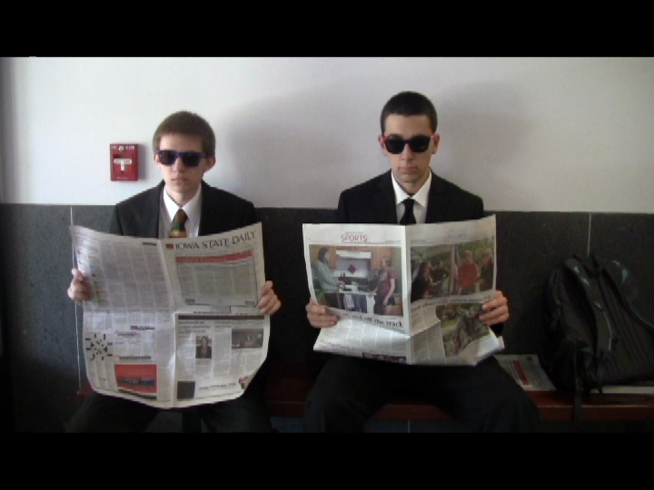 Me (left) and a friend (right) sitting on a bench in black suits and dark sunglasses. We hold newspapers.